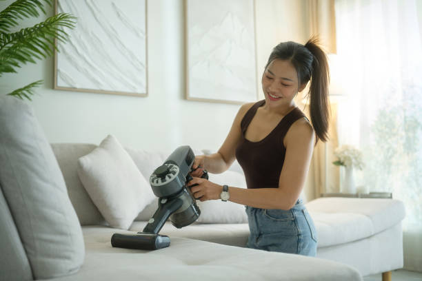 seorang wanita muda membersihkan sofa menggunakan vacuum cleaner handheld