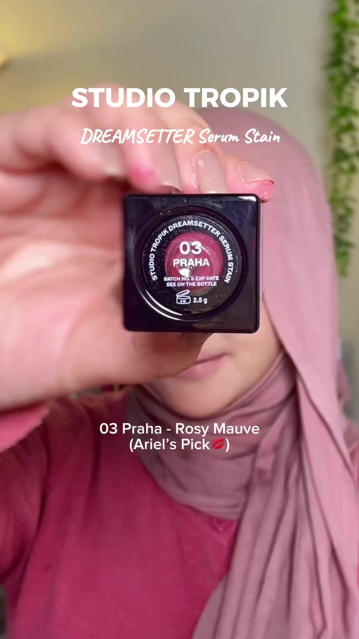 Tangan mengangkat botol Studio Tropik Dreamsetter Serum Stain menghadap kamera sehingga label shade 03 Praha terlihat, wajah tertutup botol