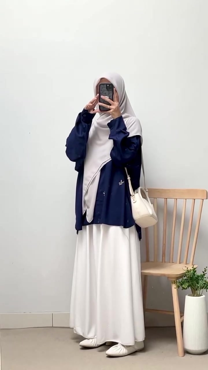 Foto cermin: perempuan berhijab abu muda memakai kemeja navy longgar, rok putih panjang, dan tas krem kecil, wajah tertutup ponsel