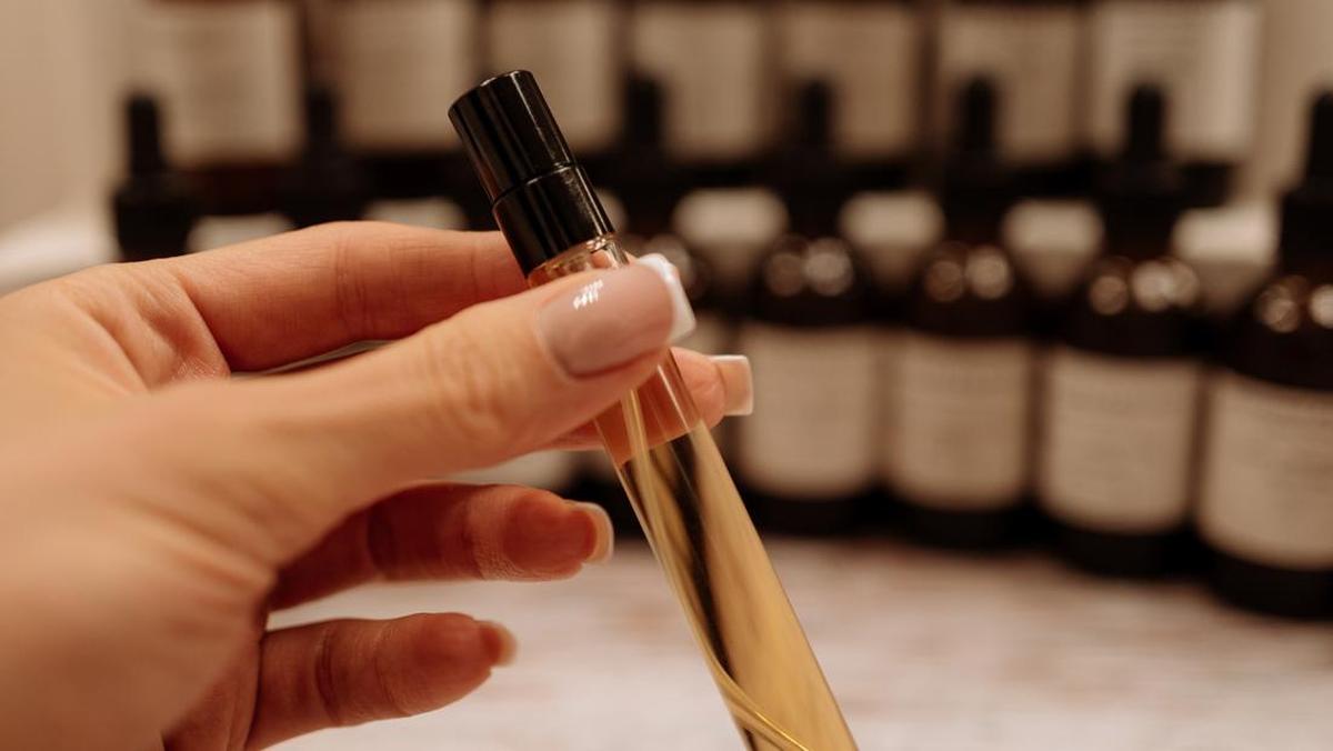 Tangan memegang vial parfum ramping berisi cairan keemasan dengan deretan botol cokelat di latar belakang