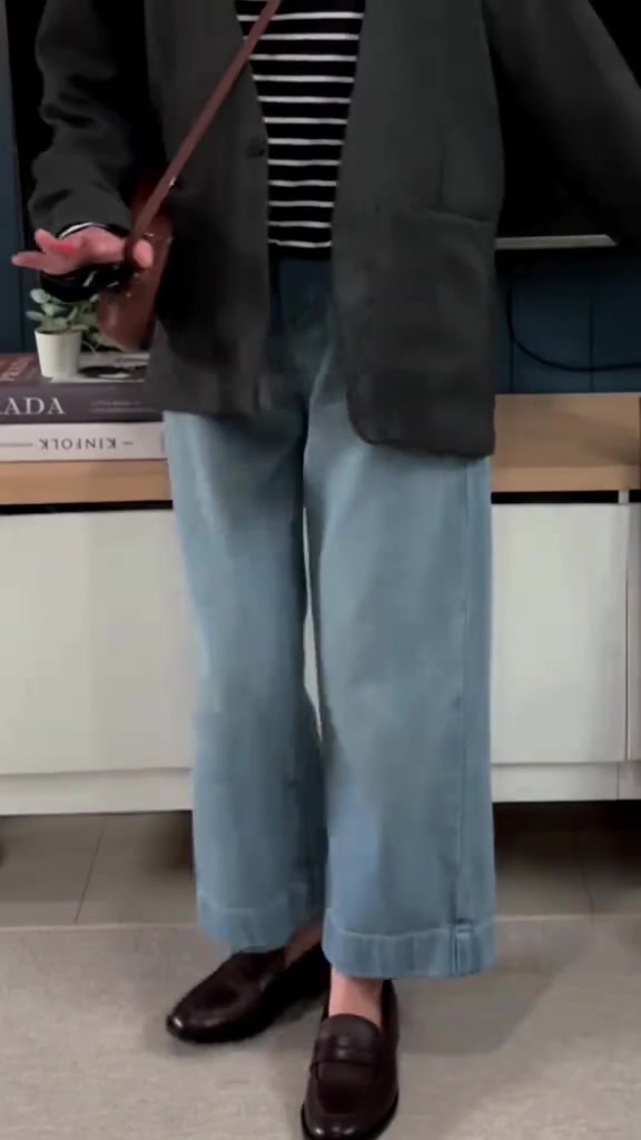 Potongan dari leher ke bawah: blazer gelap, atasan salur hitam putih, celana denim longgar biru muda, loafers kulit cokelat, dan tas crossbody
