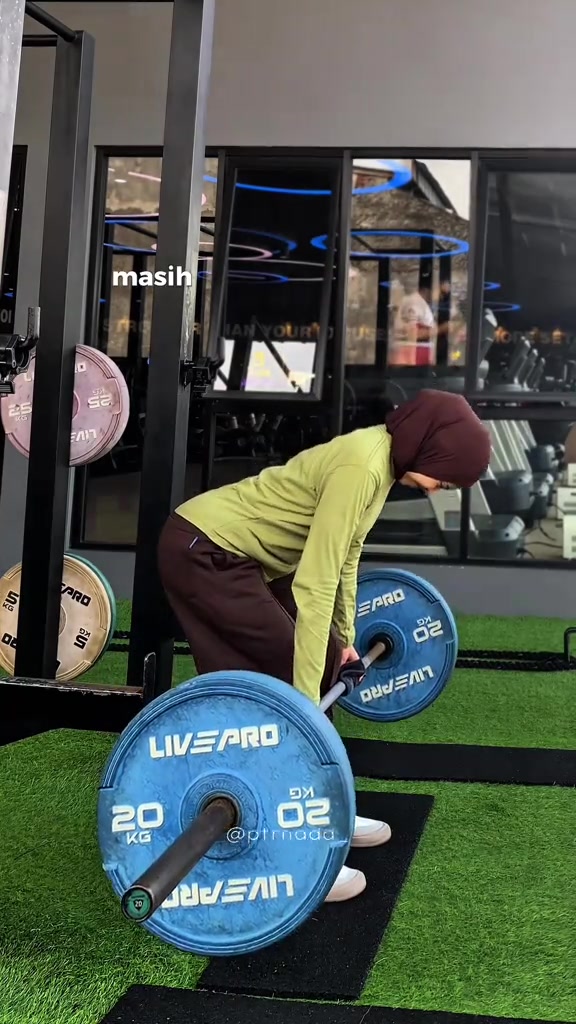 Tampak samping belakang perempuan berhijab marun beratasan longgar hijau limau membungkuk meraih barbell di gym, hem atasan tetap menutup pinggul sampai bokong