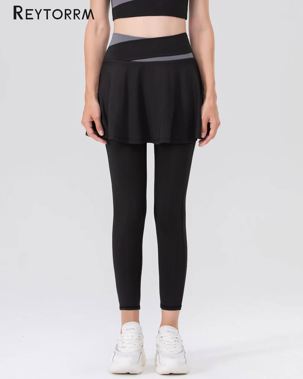 Foto produk legging olahraga hitam high waist dengan ban pinggang silang abu-abu dan rok overlay, dipakai model dari badan atas sampai sepatu di latar putih