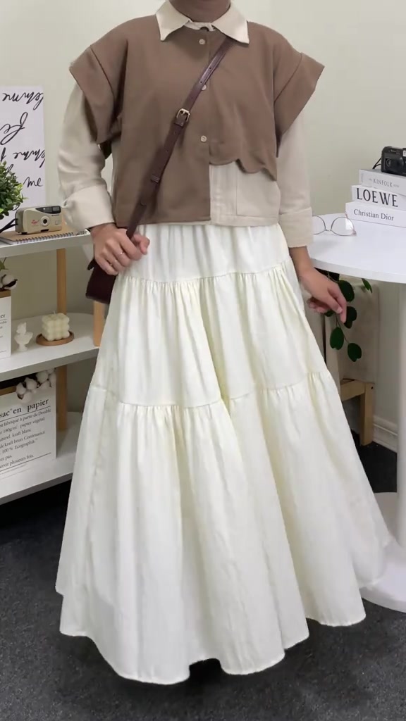Outfit dari leher ke bawah: rompi rajut cokelat di atas kemeja krem, rok putih bertingkat, dan tas selempang kecil
