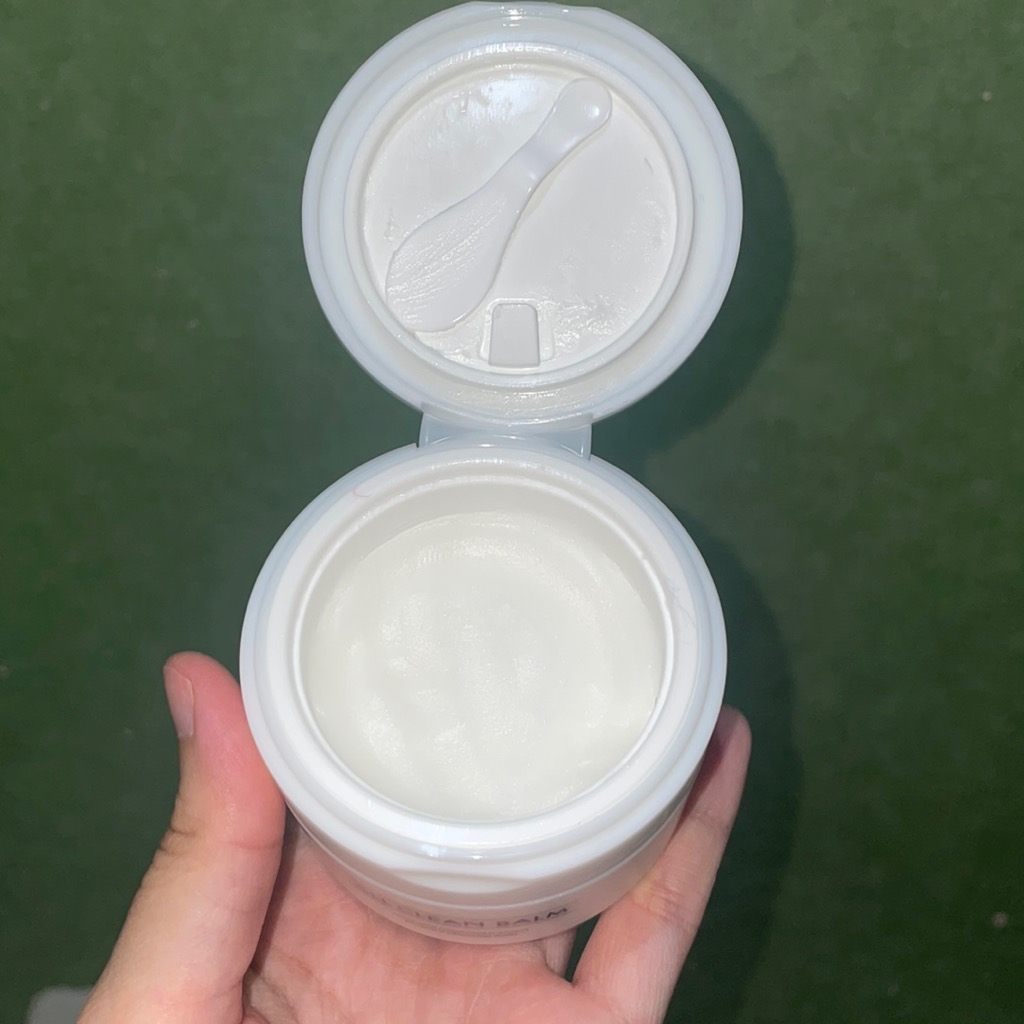 Cleansing balm putih bertekstur padat di dalam jar terbuka, lengkap dengan spatula kecil di tutup dalamnya