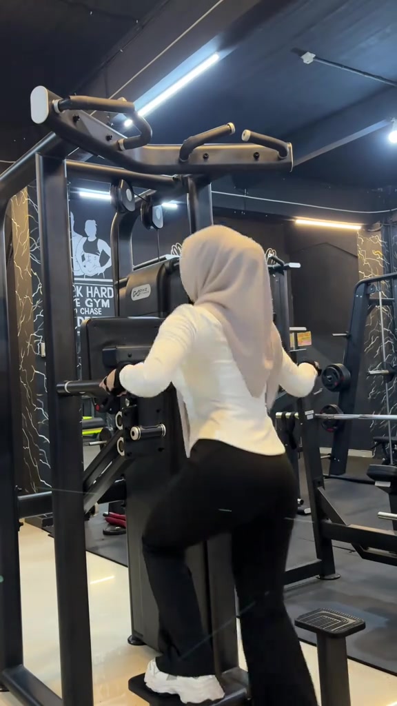 Tampak belakang perempuan berhijab krem berbaju lengan panjang putih dan celana hitam saat melangkah naik ke mesin pull-up di gym