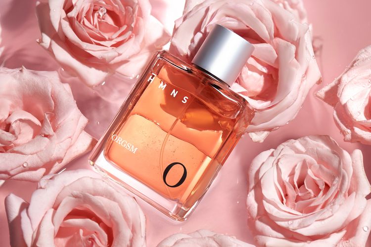 Botol parfum HMNS Orgsm berwarna oranye terbaring di antara kelopak mawar merah muda