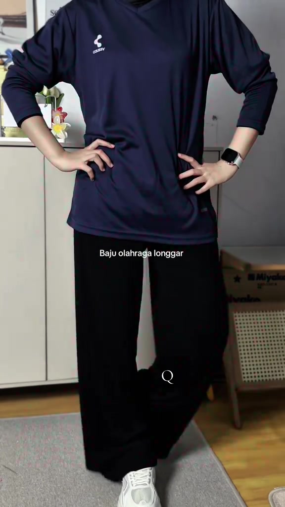 Tampak depan perempuan beratasan olahraga longgar navy berlengan panjang dengan hem melengkung dan celana hitam lebar, kedua tangan di pinggang, kepala di luar bingkai