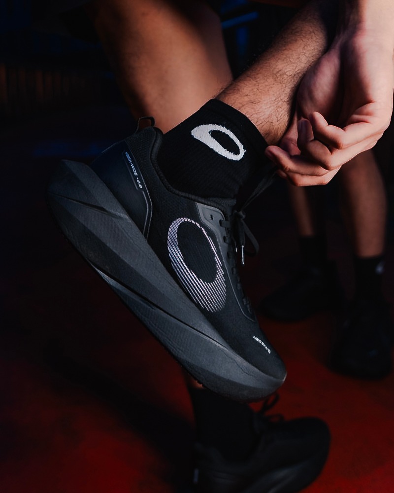 Sepatu lari Gumi Vega hitam dipakai seseorang yang sedang duduk menyesuaikan kaus kakinya