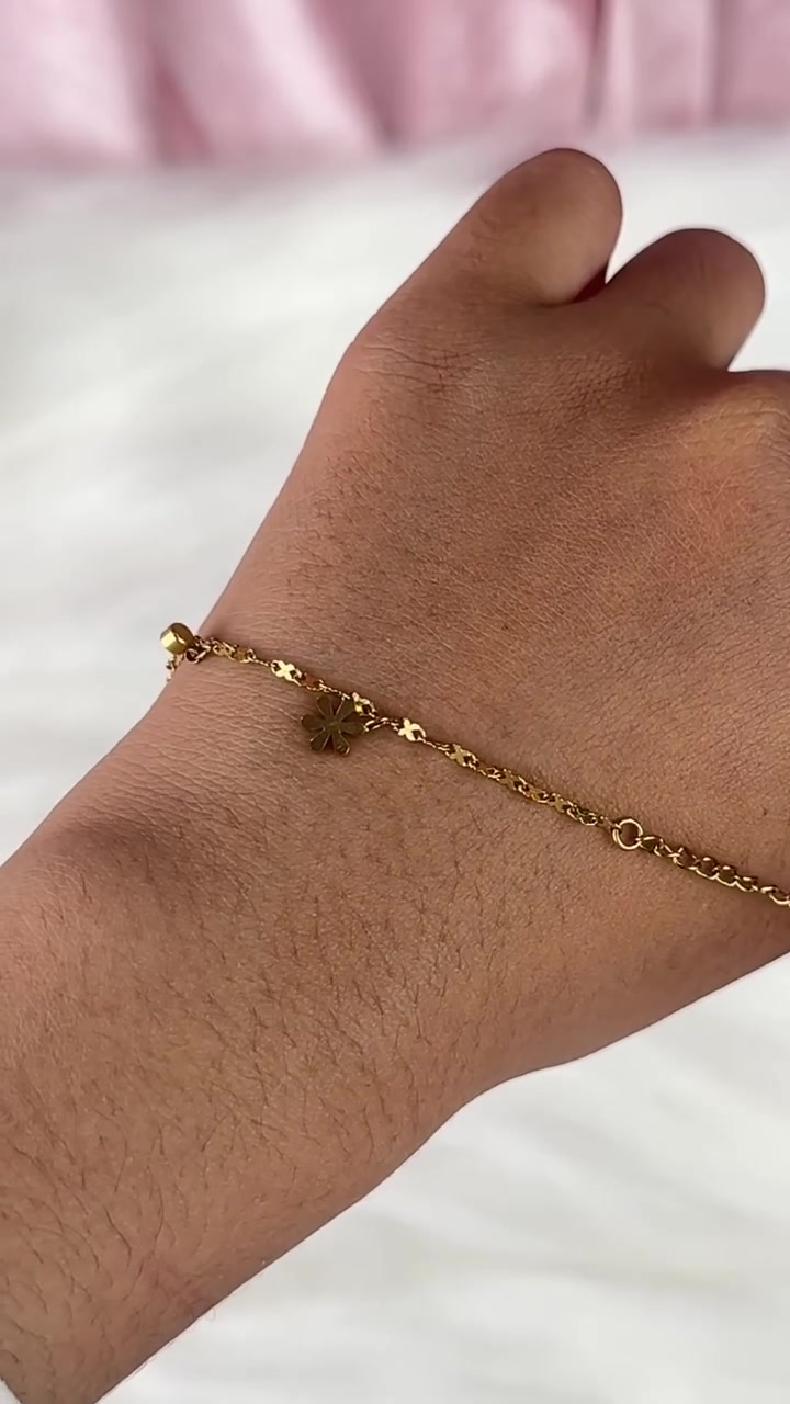 Gelang rantai emas dengan charm bunga mungil melintang di punggung tangan, latar kain putih dan pink