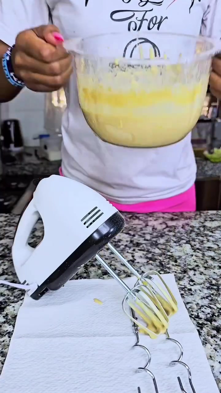 Hand mixer putih hitam dengan dua pengocok berbalut adonan kuning tergeletak di atas tisu dapur