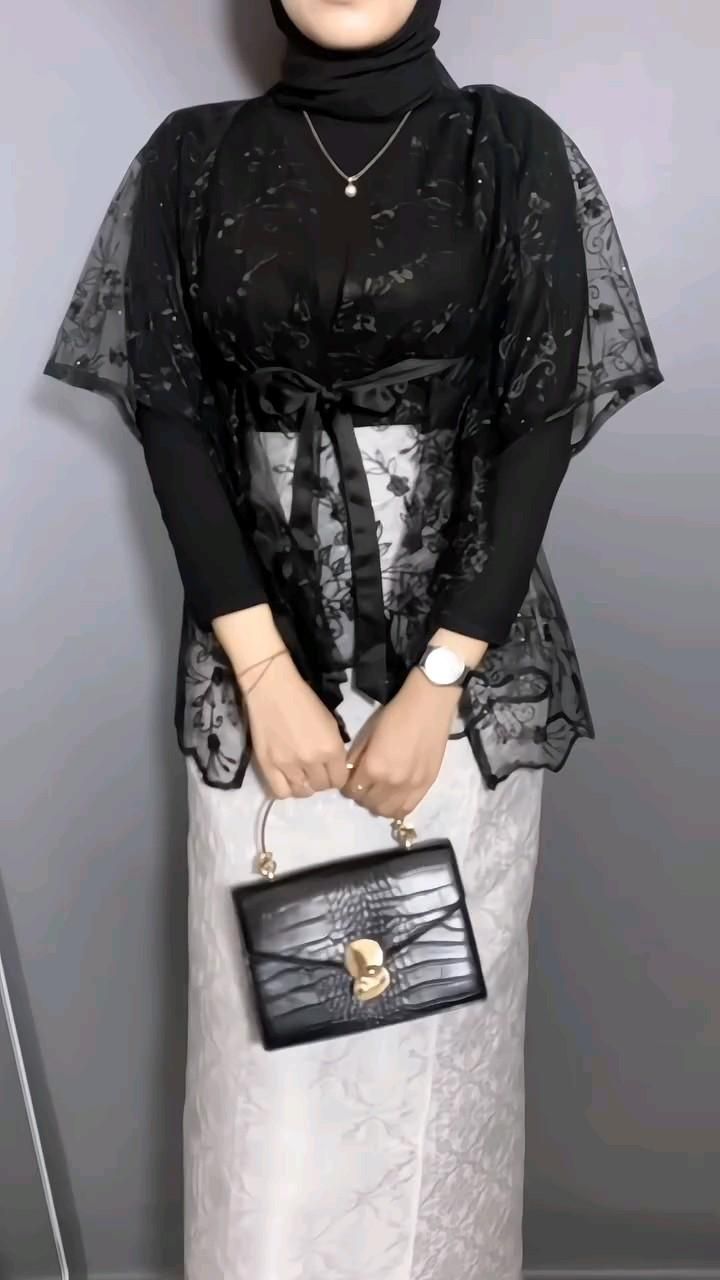 Outer brokat hitam model cape dengan ikat pinggang di atas rok putih dan tas kotak hitam, framing dari leher ke bawah