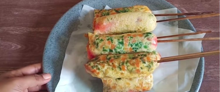Tusukan sostel kukus berbintik wortel dan daun bawang tersusun rapi di pita kertas dalam piring putih