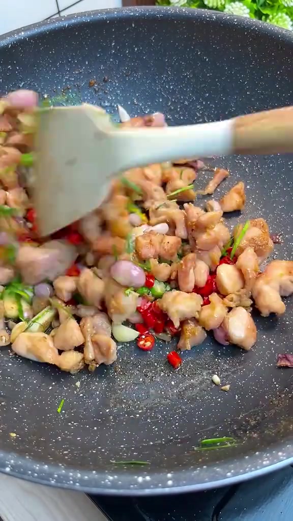 Potongan ayam oseng diaduk spatula di wajan abu-abu bersama irisan cabe merah dan serai