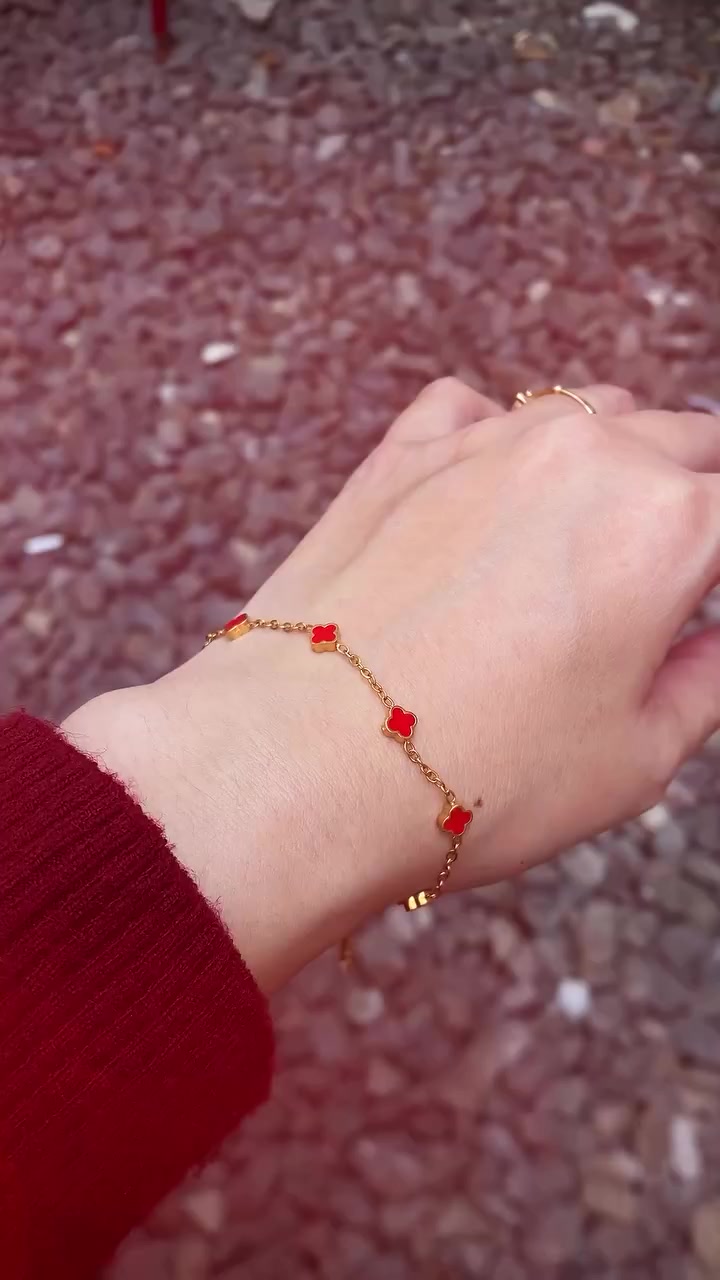 Gelang rantai emas dengan charms semanggi merah melintang di pergelangan tangan, latar kerikil halus