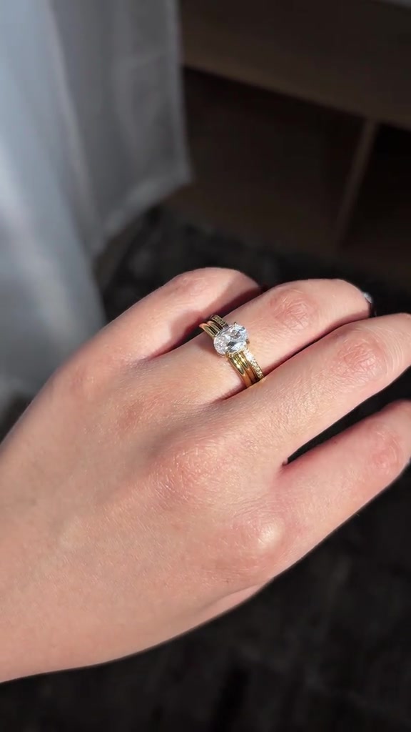 Close-up tangan di cahaya matahari memakai cincin solitaire oval yang diselipkan di tengah cincin dua layer emas dan band pavé tipis