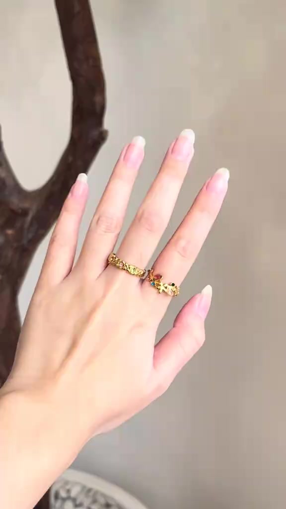 Tangan dengan dua cincin emas bertekstur di jari tengah dan jari manis dengan latar beige polos