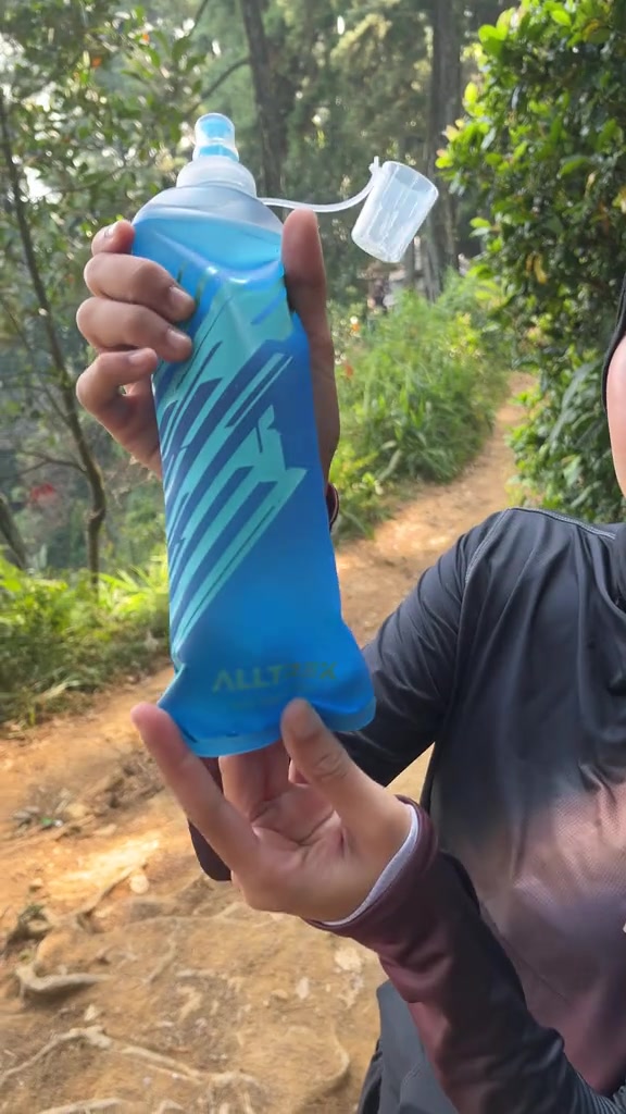 Tangan memegang softflask biru berisi air di jalur tanah berbukit