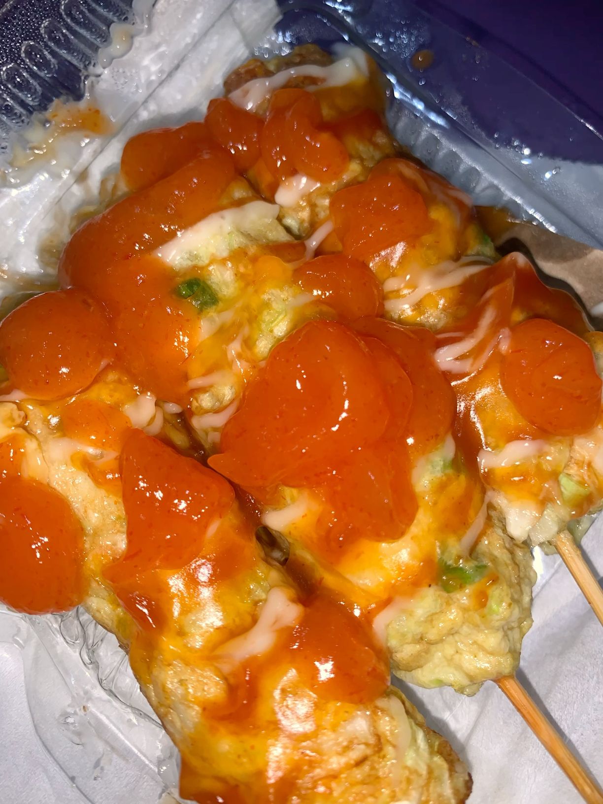 Tiga tusuk sostel goreng disiram saus sambal oranye dan garis mayones yang mengilap dalam wadah takeaway