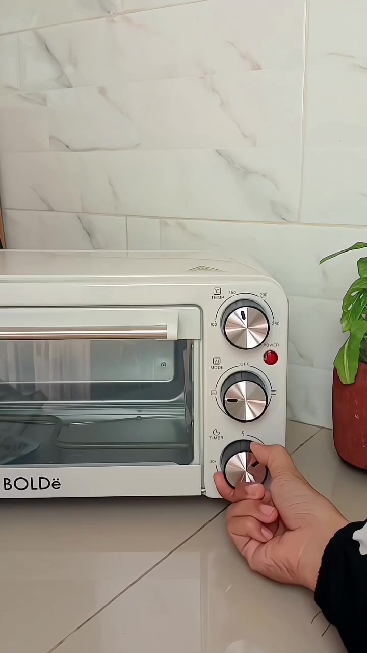 Oven listrik putih dengan tiga knob pengatur suhu, mode, dan timer yang sedang diputar tangan di dapur