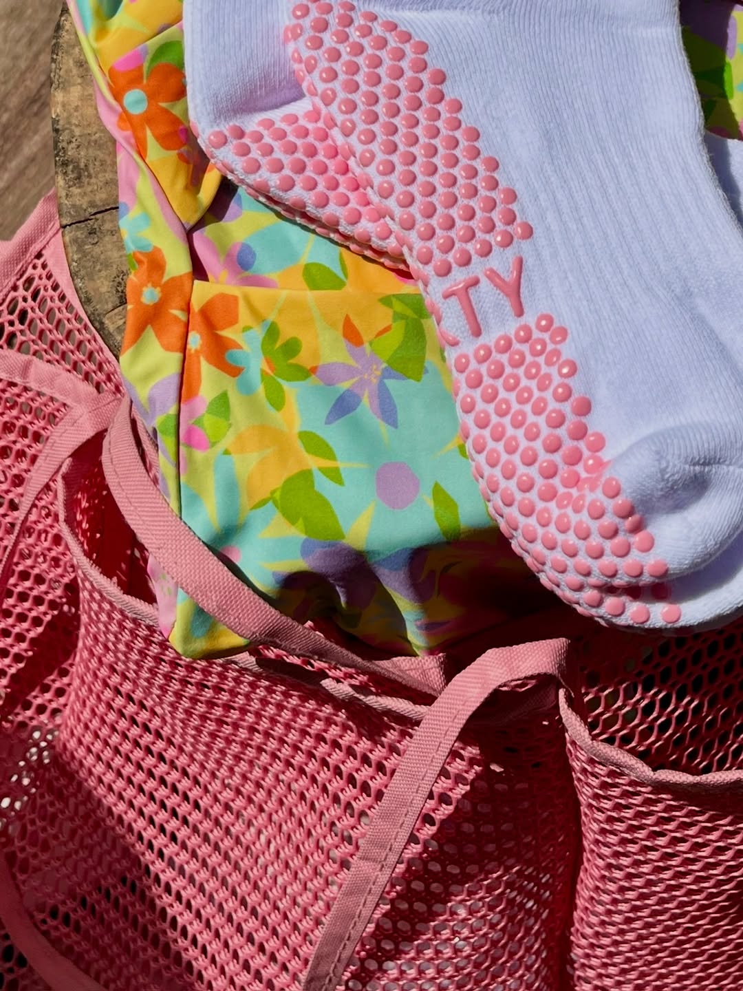 Close-up telapak grip socks putih dengan titik-titik silikon merah muda, tergeletak di atas tas jala merah muda dan kain bermotif bunga