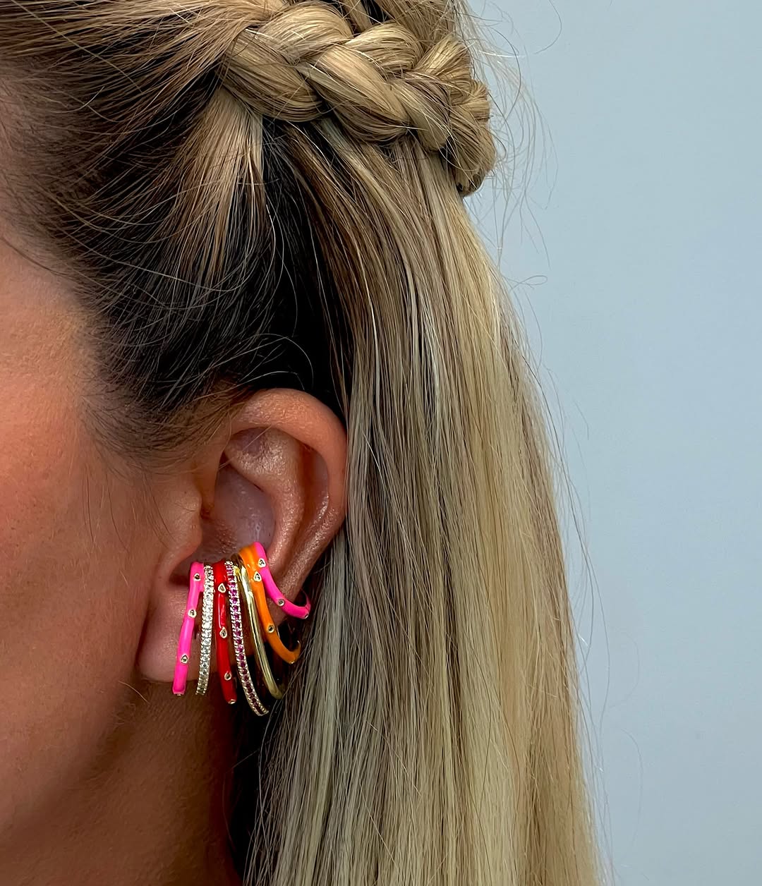 Tampak samping telinga memakai ear cuff enamel merah muda dan oranye beberapa baris, rambut pirang dikepang
