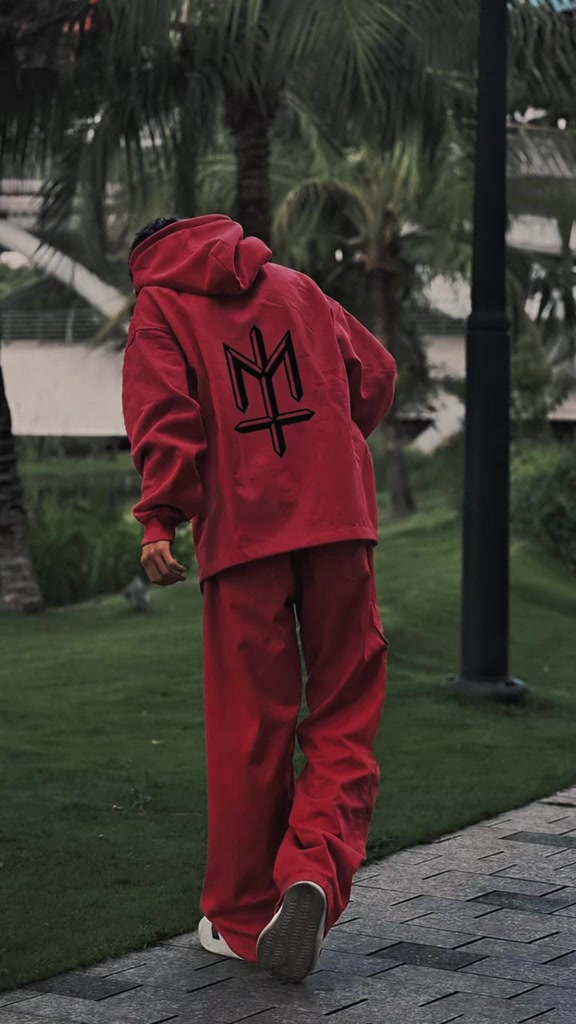 Tampak belakang seseorang berjalan di jalan setapak taman memakai setelan jas hujan merah model hoodie dengan hood dipakai