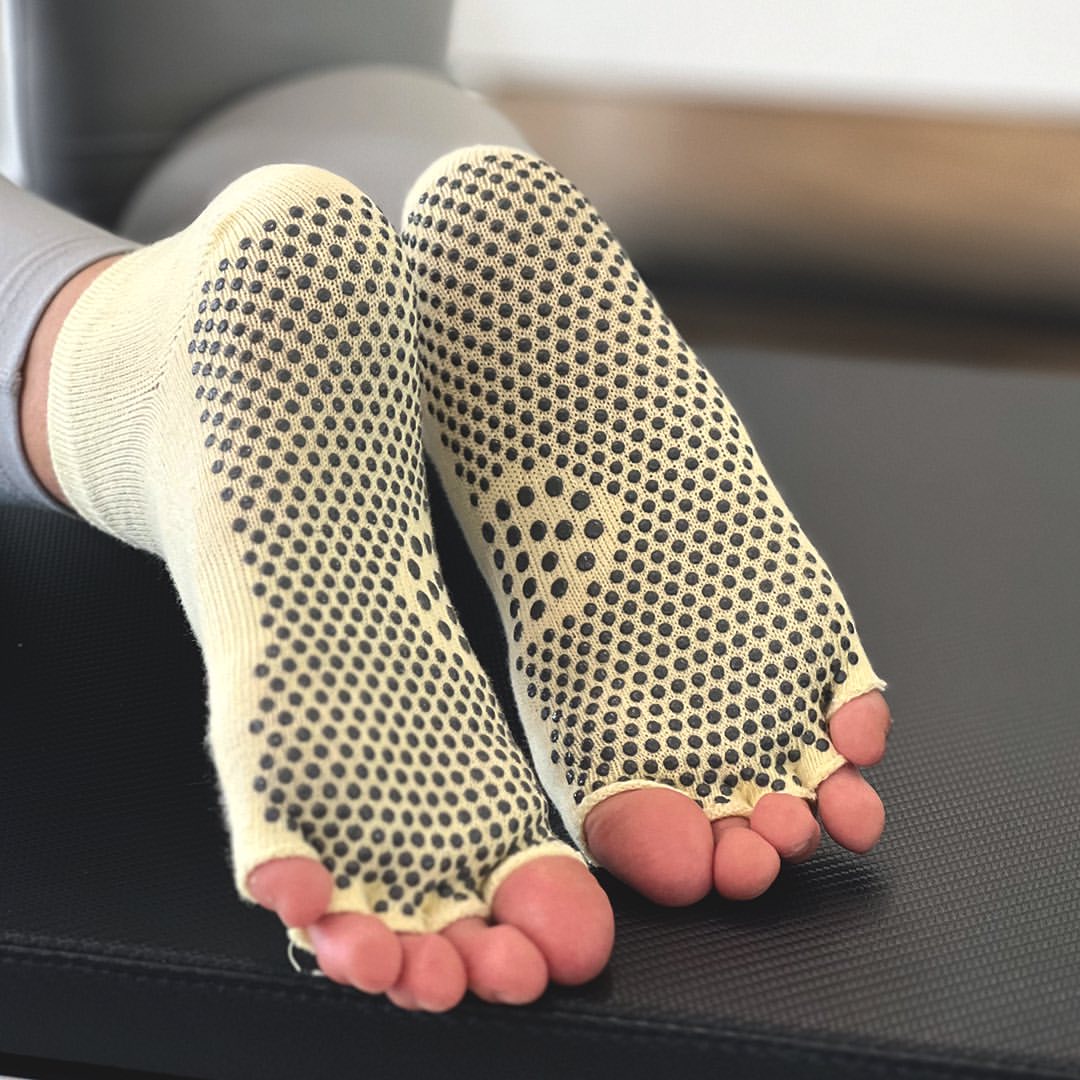 Telapak grip socks model toeless warna krem dengan grip titik hitam penuh dan jari kaki terbuka, disandarkan di permukaan alat pilates berwarna hitam