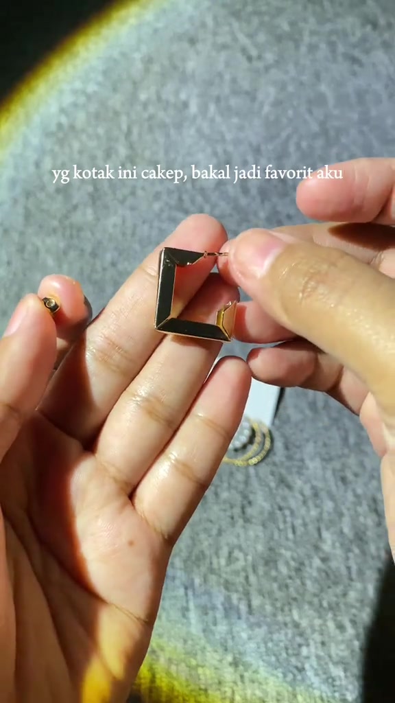 Anting klip gold bentuk kotak persegi dipegang dua jari tangan di atas latar kain abu-abu
