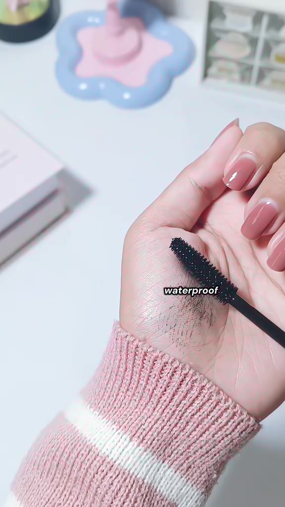 Swatch maskara hitam di telapak tangan dengan wand diletakkan di atasnya, lengan bersweater rajut pink