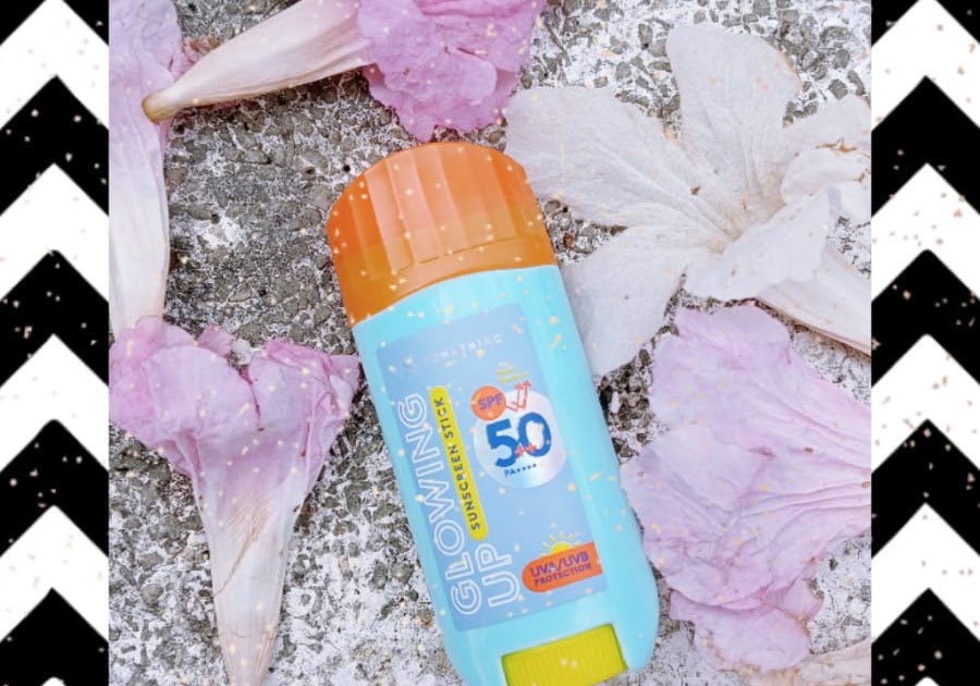 Somethinc Glowing Up Sunscreen Stick biru-oranye difoto flat lay di antara kelopak bunga putih dan pink