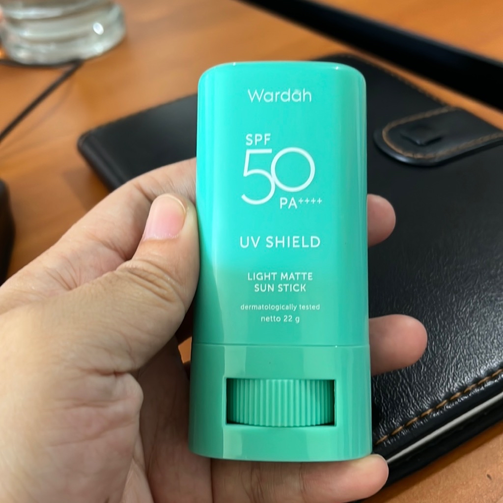 Tangan memegang sunscreen stick Wardah UV Shield Light Matte warna teal di atas meja kayu