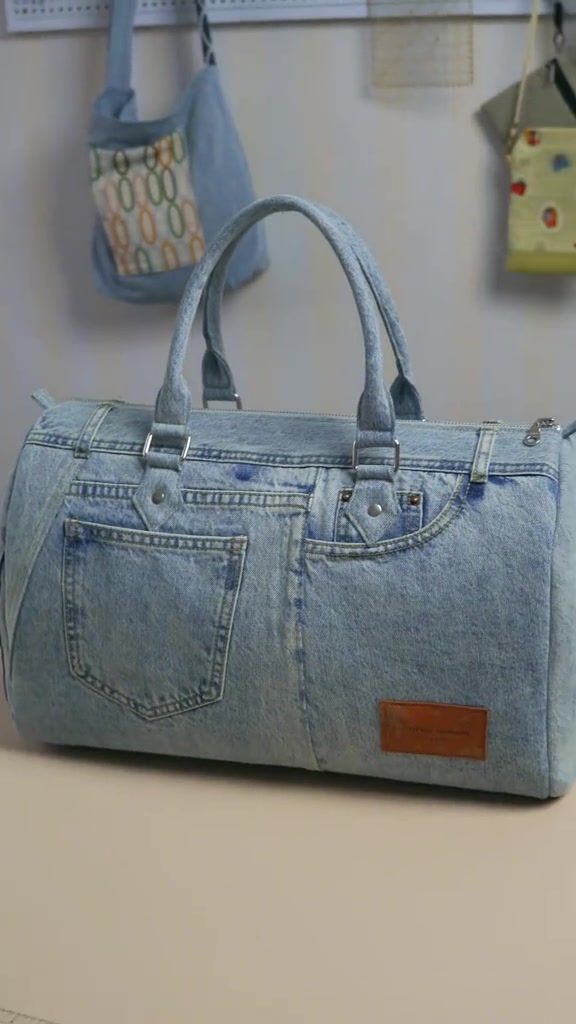 Tas boston denim jahitan dari jeans bekas dengan dua handle, kantong belakang, dan patch kulit, diletakkan di meja kerja