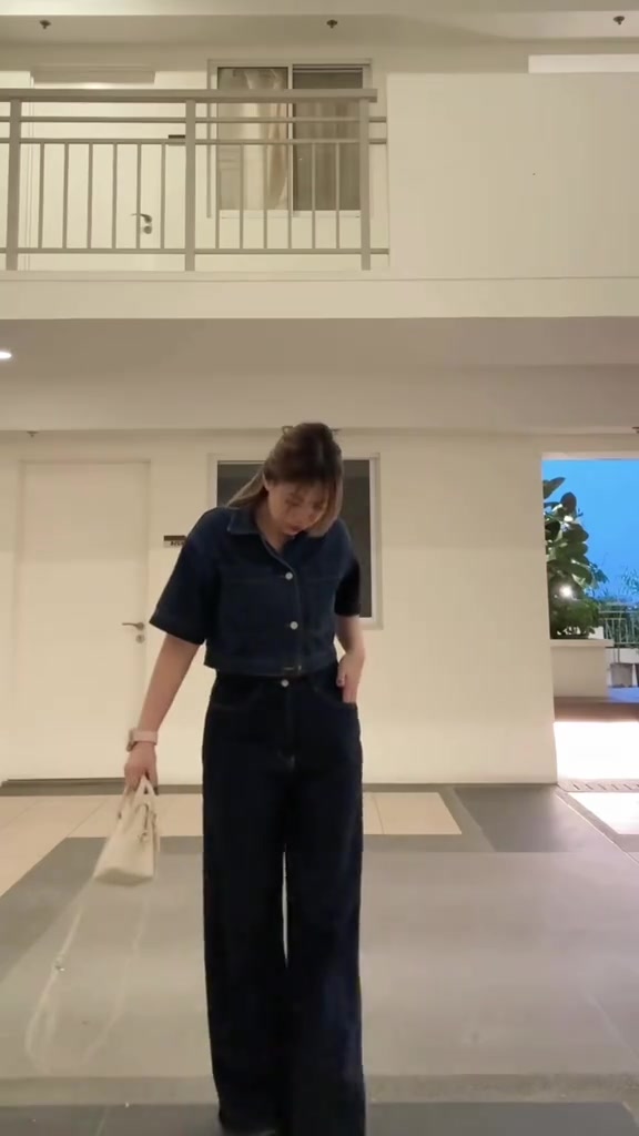 Perempuan menunduk memakai kemeja denim pendek dan celana denim gelap longgar sambil menjinjing tas kecil warna krem di lorong terbuka