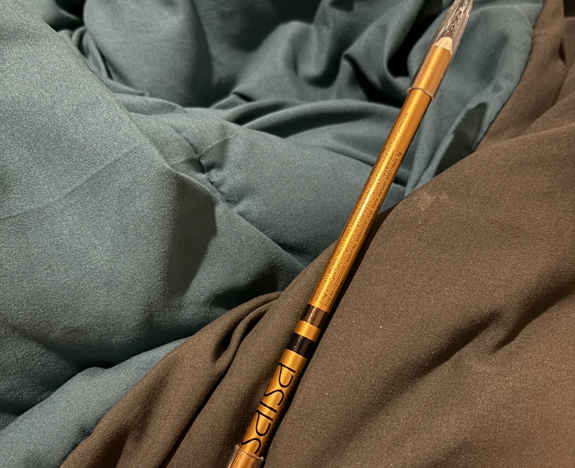 Pensil alis batang emas tergeletak diagonal di atas kain abu-abu dan cokelat