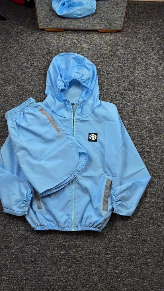 Set jaket dan celana parasut running warna biru muda terbentang di lantai karpet, lengkap dengan hood dan strip reflektif
