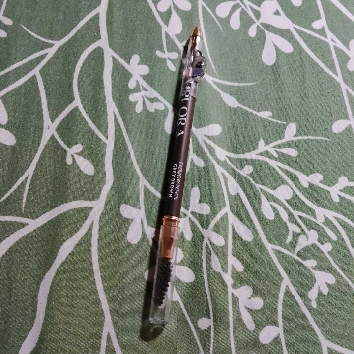 Pensil alis Implora Grey Brown batang cokelat dengan tutup spoolie, tergeletak di atas kain hijau motif daun