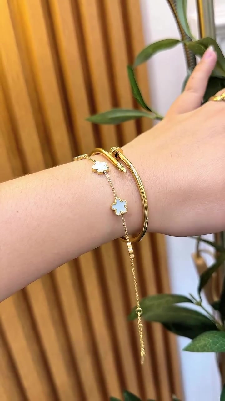 Pergelangan tangan memakai gelang rantai clover putih dan bangle emas dengan latar tanaman hijau