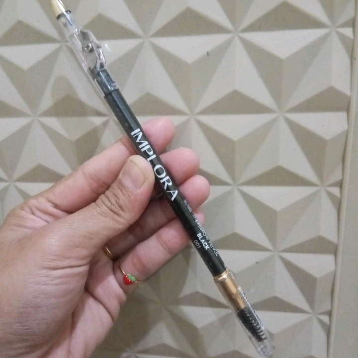 Pensil alis Implora hitam 2in1 dengan tutup spoolie transparan, dipegang di depan latar dinding bertekstur