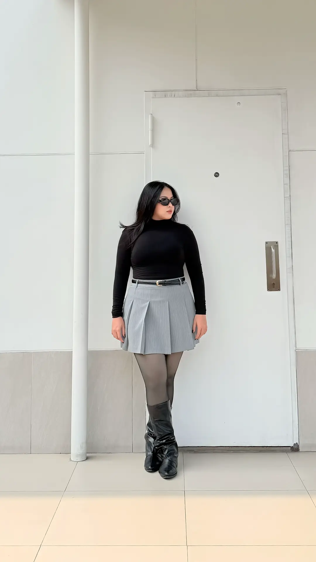 Atasan turtleneck hitam fitted dipadu rok plisket mini abu-abu, belt kulit tipis, tights gelap, dan boots hitam di depan dinding putih