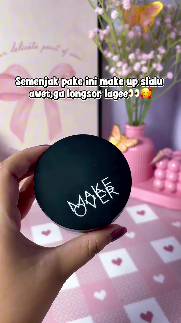 Tangan memegang jar Make Over Silky Smooth Translucent Powder tutup hitam berlogo putih dengan latar meja merah muda