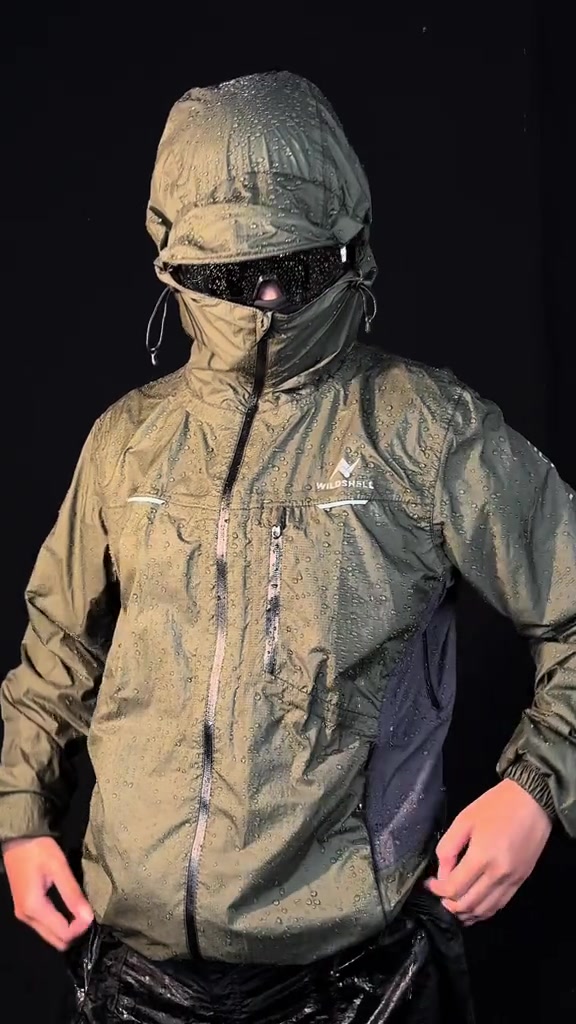 Jaket hujan hijau zaitun berhood dengan butiran air memenuhi permukaan kain, wajah pemakainya tertutup masker dan goggle