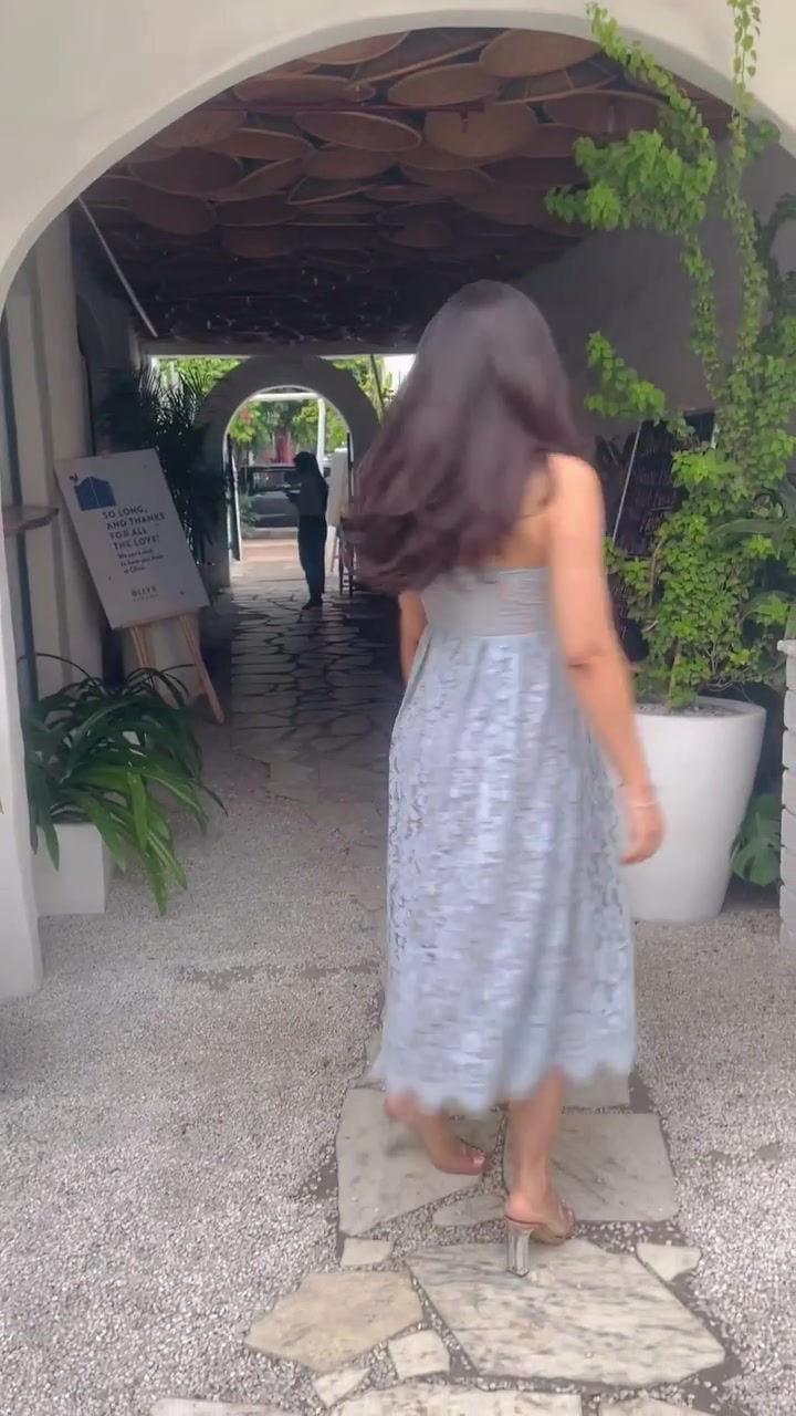 Tampak belakang perempuan berjalan di lorong beratap lengkung memakai dress midi lace biru keabuan dengan hem scalloped, heels transparan, dan tas oranye