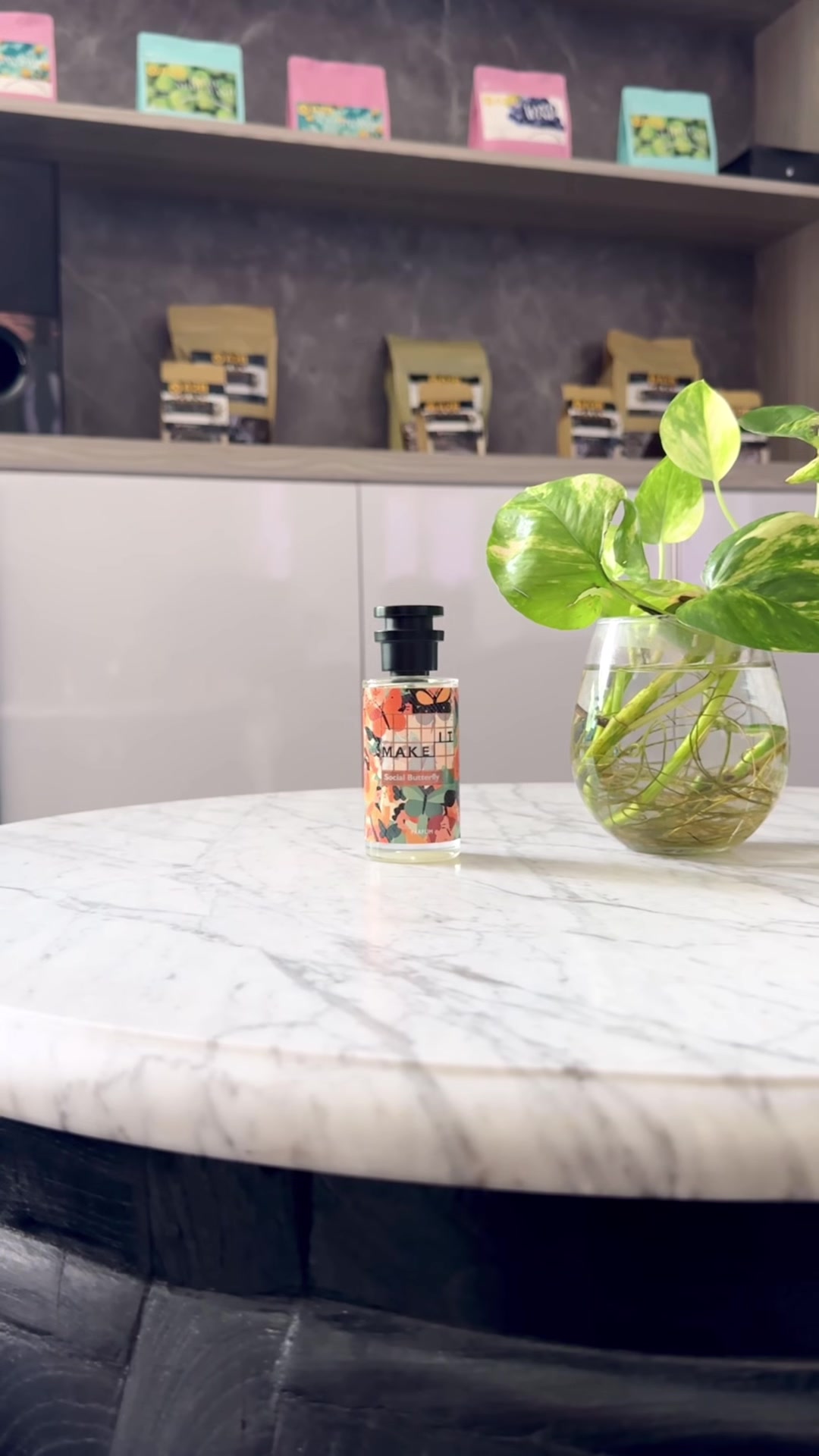 Botol Make It Social Butterfly berlabel biru muda di meja marmer samping vas kaca berisi tanaman di kafe