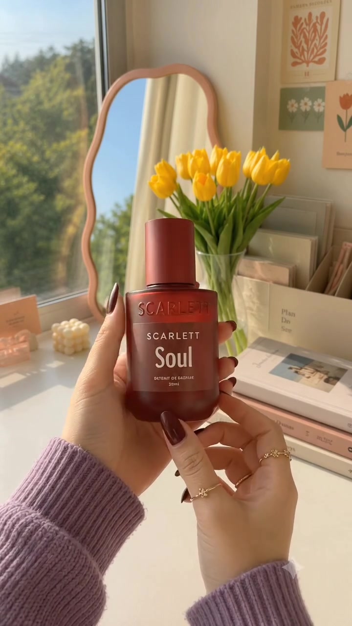 Dua tangan memegang botol Scarlett X Soul merah marun berlabel putih di depan vas tulip kuning, cermin bergelombang, dan tumpukan buku pada meja putih dekat jendela yang cerah