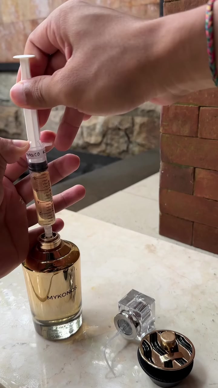 Tangan memegang spuit yang menyedot juice dari botol emas Mykonos Monaco Royale di atas meja marmer luar ruangan, dengan atomizer dan tutup botol tergeletak di sampingnya