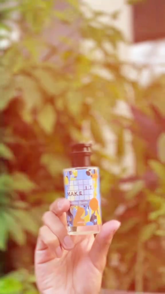 Tangan mengangkat botol Make It Weekday Hustle berlabel biru oranye dengan latar dedaunan hangat yang kabur