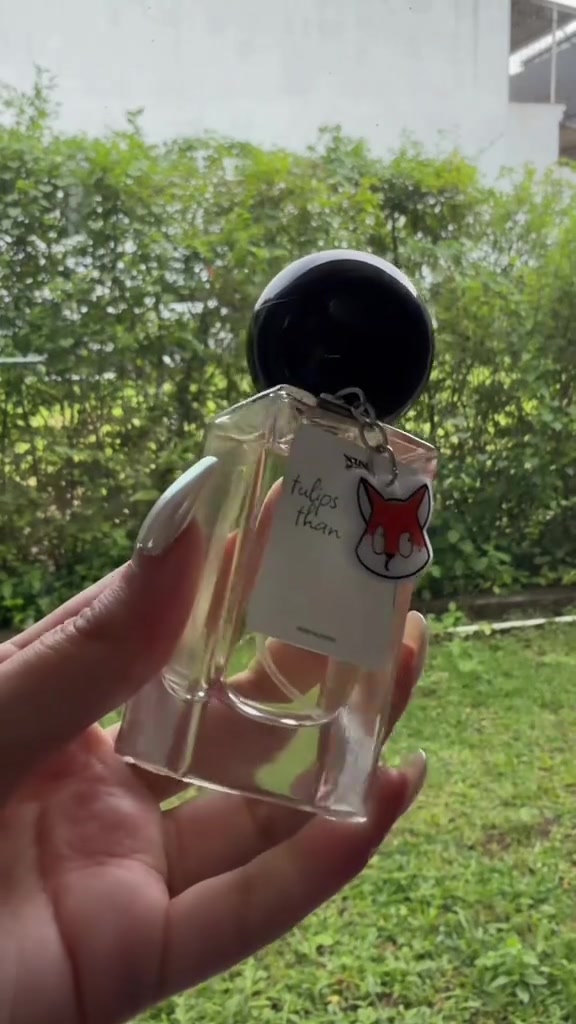 Tangan memegang botol Scuse Me Perfumery Tulips Better Than One berlabel putih dengan gantungan rubah kecil di depan pagar tanaman hijau