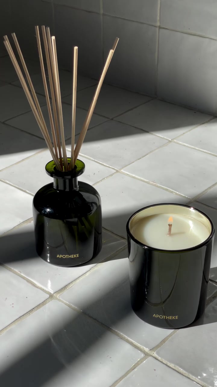 Reed diffuser botol kaca gelap dengan stik kayu di samping lilin menyala di atas lantai keramik putih terkena cahaya matahari
