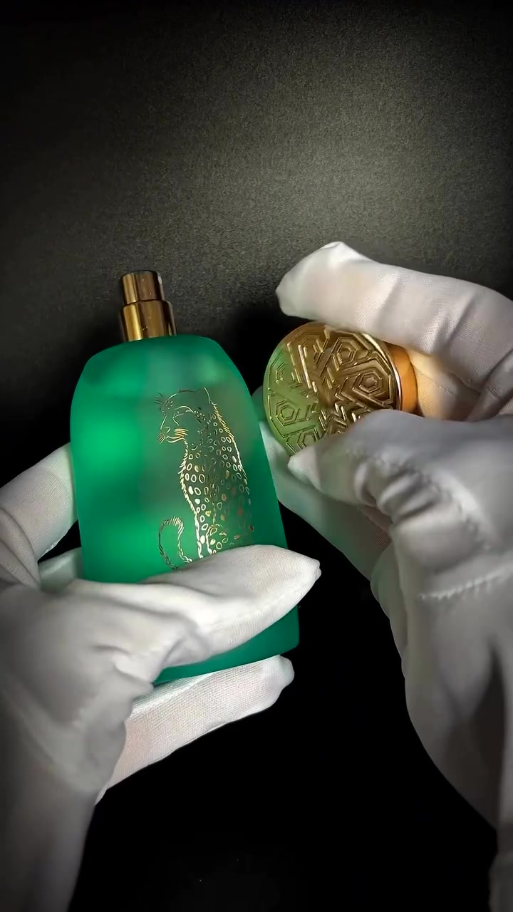 Dua tangan bersarung putih memegang botol Mykonos Dreamscape hijau tosca dalam keadaan terbuka beserta tutup emas berukir di depan latar gelap
